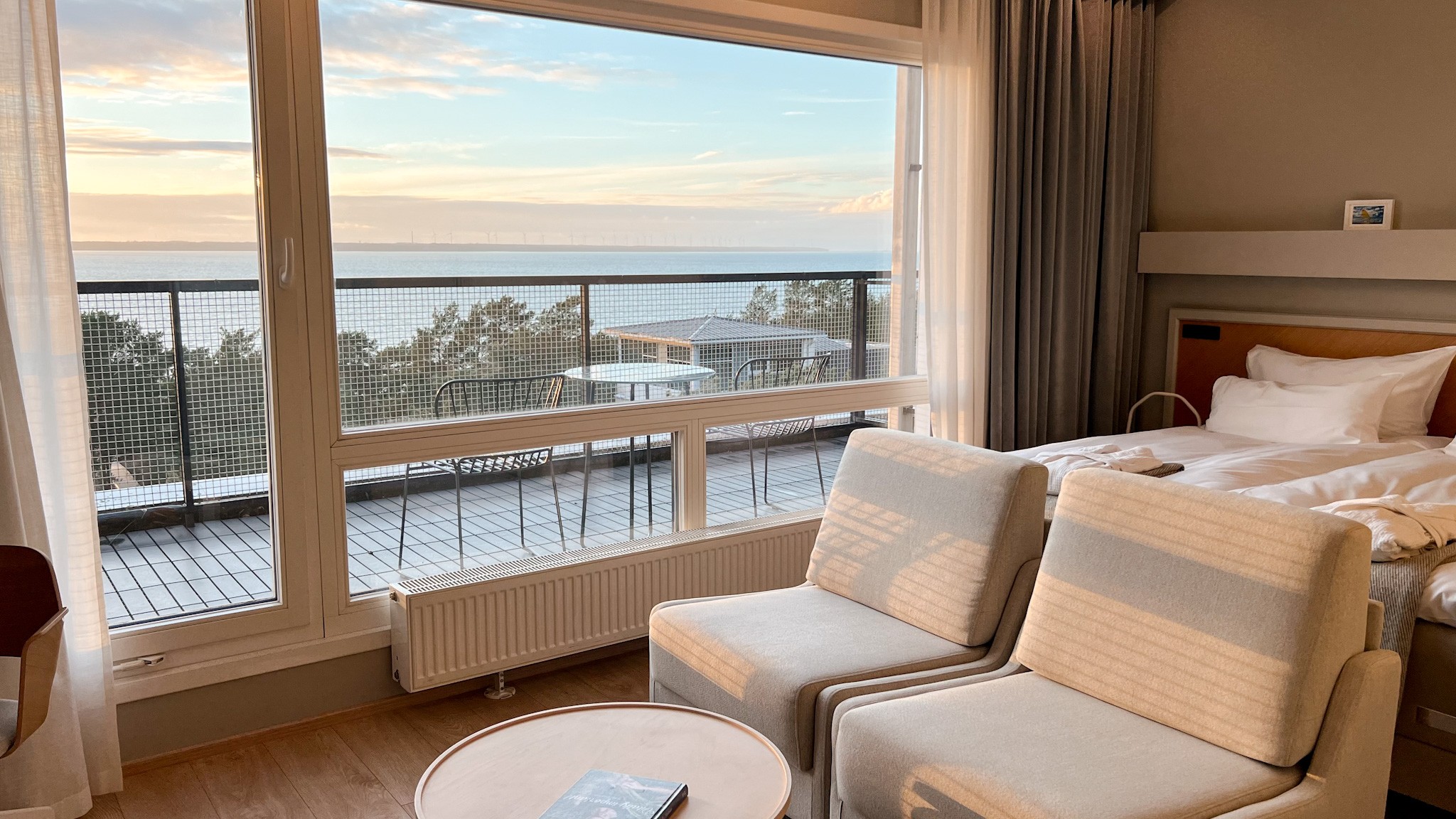 Hotel room with large windows and balcony overlooking the sea, soft morning light, minimal Nordic interior with bed and lounge chairs